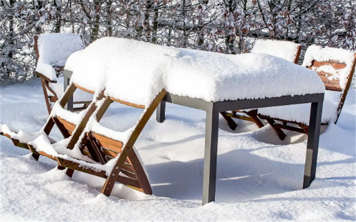 Unprotected wooden outdoor table and lounge chairs blanketed with heavy winter snow in cold climate backyard